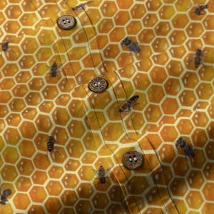 May include: Close-up of a yellow and orange patterned fabric with a honeycomb design and scattered bees. The fabric has three wooden buttons. The design is a repeating pattern of hexagonal shapes and bees.