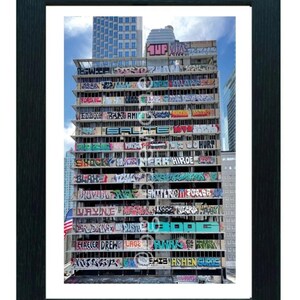 May include: A framed print of a multi-story building covered in colorful graffiti art. The artwork includes various words and phrases, with an American flag visible on the side. The image is set against a backdrop of a blue sky and other buildings.
