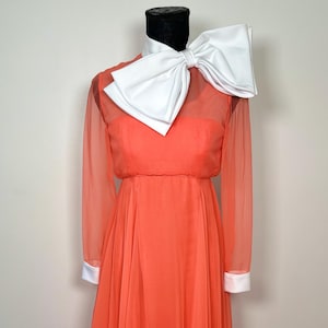 May include: Coral-colored formal gown with a large white bow collar. The long-sleeved dress features white cuffs and a flowing skirt. The dress is displayed on a mannequin.