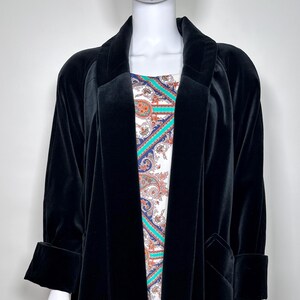 May include: A long, black velvet coat with a wide lapel and cuffed sleeves. The coat is open, revealing a colorful patterned dress underneath. The dress features a paisley design with shades of blue, orange, and white.