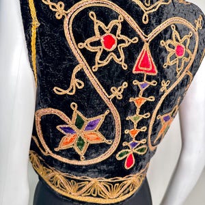 Puede incluir: Un chaleco de terciopelo negro con intrincados bordados en hilo dorado y aplicaciones coloridas en forma de estrella en rojo, verde, naranja y morado. La prenda incluye un borde decorativo dorado a lo largo del dobladillo y las sisas, mostrando patrones de costura detallados.
