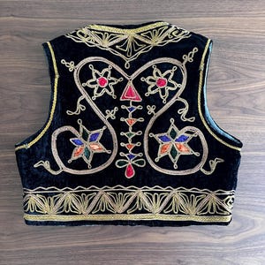 Puede incluir: Un chaleco de terciopelo negro con intrincados bordados met&aacute;licos dorados a lo largo de los bordes y el escote. El panel trasero muestra un dise&ntilde;o sim&eacute;trico con motivos coloridos en forma de estrella en rojo, azul, verde y naranja.