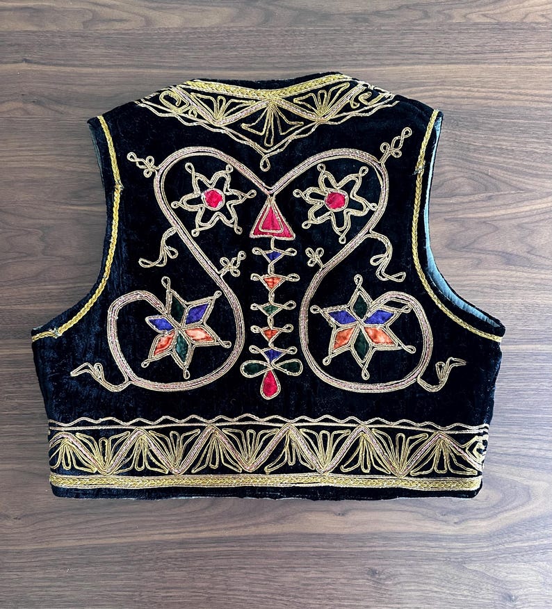Puede incluir: Un chaleco de terciopelo negro con intrincados bordados met&aacute;licos dorados a lo largo de los bordes y el escote. El panel trasero muestra un dise&ntilde;o sim&eacute;trico con motivos coloridos en forma de estrella en rojo, azul, verde y naranja.