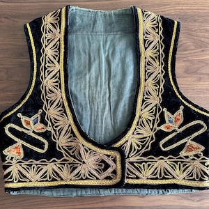 Puede incluir: Un chaleco de terciopelo negro con intrincados bordados en hilo dorado a lo largo de los bordes delanteros y el dobladillo. La prenda incluye motivos decorativos florales en naranja y azul en los paneles del pecho y forro verde claro.