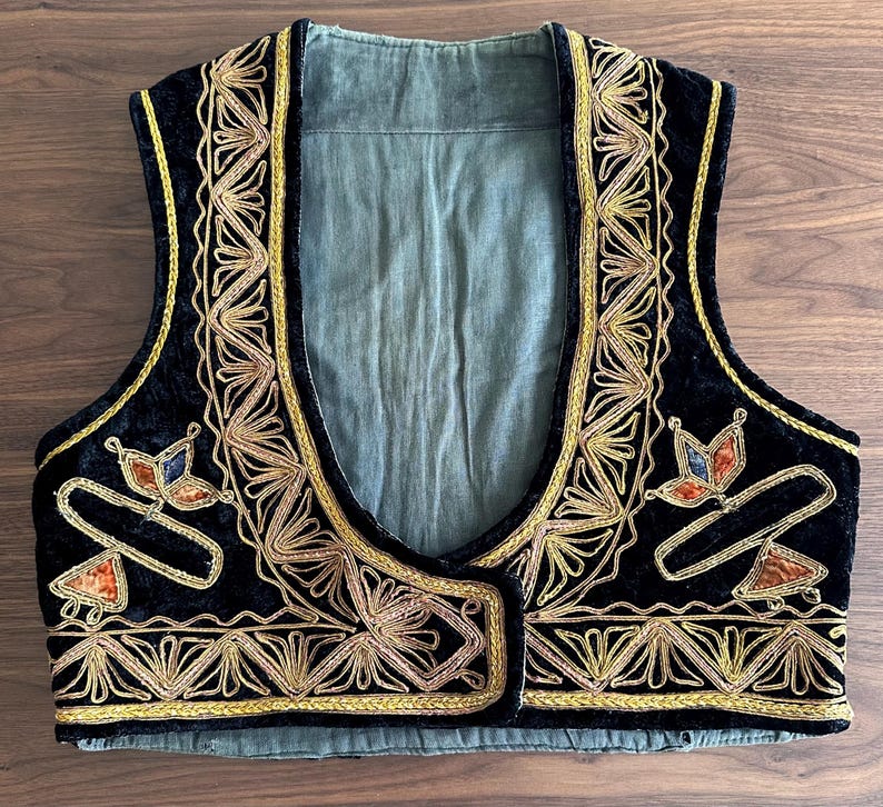 Puede incluir: Un chaleco de terciopelo negro con intrincados bordados en hilo dorado a lo largo de los bordes delanteros y el dobladillo. La prenda incluye motivos decorativos florales en naranja y azul en los paneles del pecho y forro verde claro.