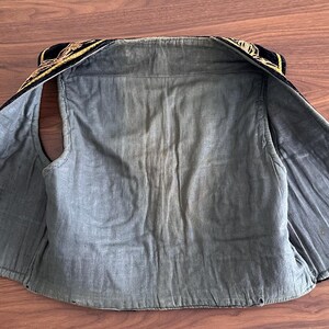 Puede incluir: Un chaleco sin mangas con forro interior azul oscuro y exterior negro adornado con intrincados bordados dorados a lo largo del cuello y los hombros. La prenda est&aacute; extendida en plano, mostrando su confecci&oacute;n, costuras y dise&ntilde;o abierto.