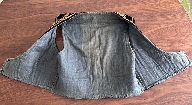 Puede incluir: Un chaleco sin mangas con forro interior azul oscuro y exterior negro adornado con intrincados bordados dorados a lo largo del cuello y los hombros. La prenda est&aacute; extendida en plano, mostrando su confecci&oacute;n, costuras y dise&ntilde;o abierto.