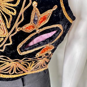 Puede incluir: Un chaleco de terciopelo negro con intrincados bordados de cord&oacute;n dorado y motivos florales decorativos. La prenda incluye un detalle de corte distintivo que revela un forro de tela rosa suave, con ribete trenzado dorado en la sisa y el dobladillo.