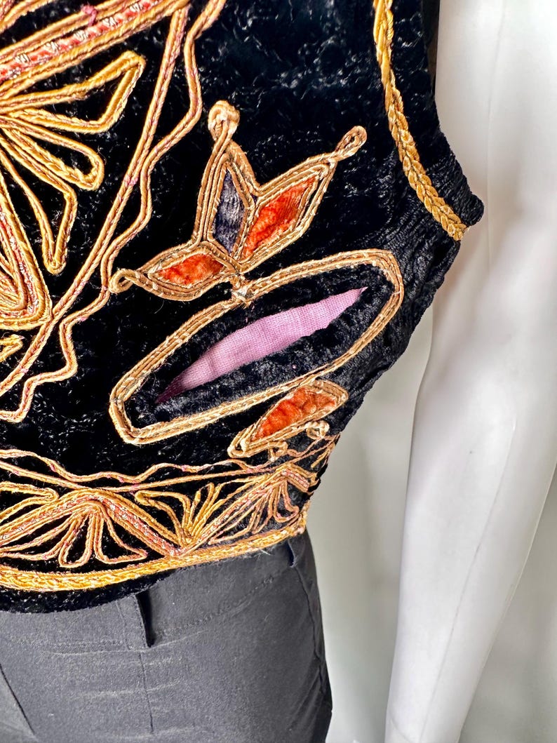 Puede incluir: Un chaleco de terciopelo negro con intrincados bordados de cord&oacute;n dorado y motivos florales decorativos. La prenda incluye un detalle de corte distintivo que revela un forro de tela rosa suave, con ribete trenzado dorado en la sisa y el dobladillo.
