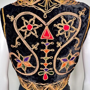 Puede incluir: Un chaleco de terciopelo negro con intrincados bordados de hilo dorado en patrones de remolinos. El dise&ntilde;o incluye motivos en forma de estrella con acentos de tela roja, morada, naranja y verde, junto con una disposici&oacute;n vertical central de formas geom&eacute;tricas.