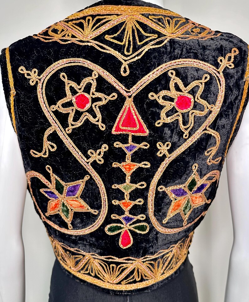 Puede incluir: Un chaleco de terciopelo negro con intrincados bordados de hilo dorado en patrones de remolinos. El dise&ntilde;o incluye motivos en forma de estrella con acentos de tela roja, morada, naranja y verde, junto con una disposici&oacute;n vertical central de formas geom&eacute;tricas.