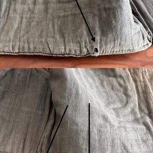 Puede incluir: Una prenda de tela texturizada de color topo con costuras visibles a lo largo de los bordes. El material muestra una apariencia tejida similar al lino con tres peque&ntilde;os agujeros circulares oscuros resaltados por flechas negras que se&ntilde;alan las &aacute;reas.