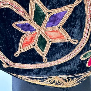 Puede incluir: Una prenda de terciopelo negro con intrincados bordados de hilo met&aacute;lico dorado y aplicaciones en forma de estrella. El dise&ntilde;o de estrella incorpora inserciones de tela en tonos vibrantes morado, verde y naranja, enmarcadas por un cord&oacute;n dorado.