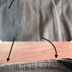 Puede incluir: Una vista de cerca de una prenda de tela tejida de color gris oscuro con un dobladillo cosido. La imagen incluye flechas negras que se&ntilde;alan &aacute;reas espec&iacute;ficas a lo largo del borde superior de la tela, resaltando la textura y los detalles de costura.
