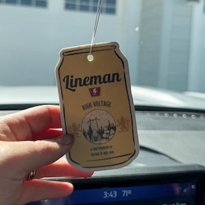 May include: Tan rectangular air freshener with the text "Lineman High Voltage" and a graphic of a power line and trees. The air freshener is hanging from a string in a car.