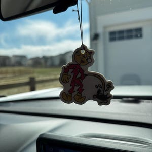 May include: A novelty air freshener shaped like a cartoon character in a red outfit, with a yellow face and shoes. The air freshener is hanging from a string in front of a car's dashboard, with a blurred outdoor background.
