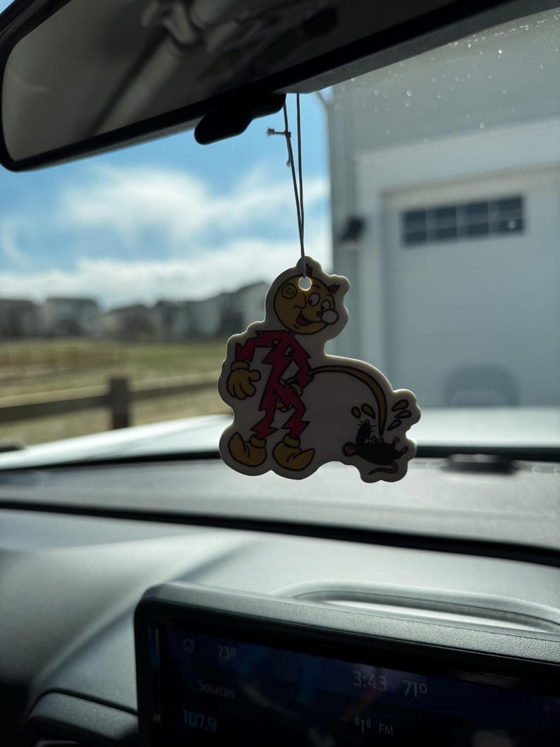 May include: A novelty air freshener shaped like a cartoon character in a red outfit, with a yellow face and shoes. The air freshener is hanging from a string in front of a car's dashboard, with a blurred outdoor background.