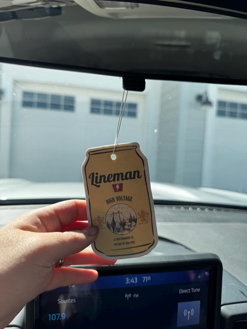 May include: Tan rectangular air freshener with the text "Lineman High Voltage" and a graphic of a power line and trees. The air freshener is hanging from a string in a car.