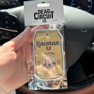 May include: An air freshener in a clear plastic package. The air freshener is shaped like a beer can with the word "Lineman" and "High Voltage" printed on it. The packaging also has the words "Dead Circuit Co." printed on it.