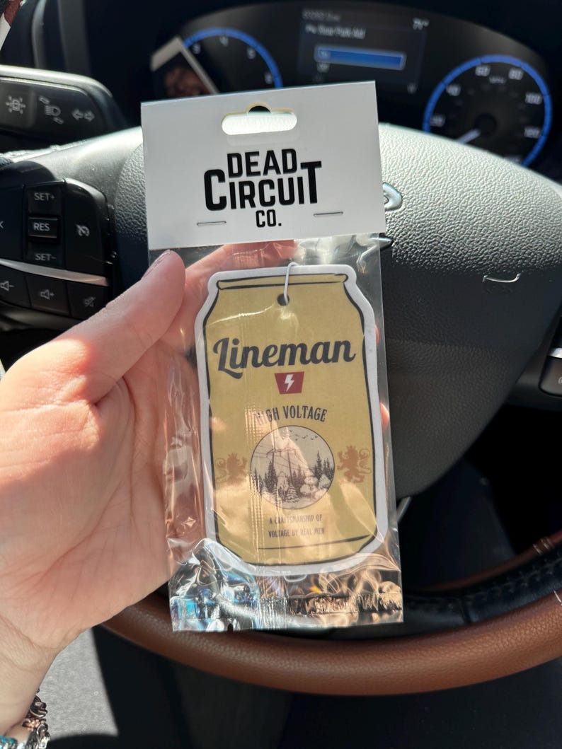 May include: An air freshener in a clear plastic package. The air freshener is shaped like a beer can with the word "Lineman" and "High Voltage" printed on it. The packaging also has the words "Dead Circuit Co." printed on it.