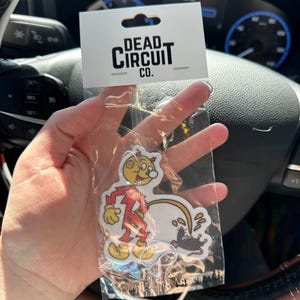 May include: A clear plastic package containing a sticker with a cartoon character in a red jumpsuit and a yellow face. The sticker also features a black rat and the text "DEAD CIRCUIT CO." on a white tag.