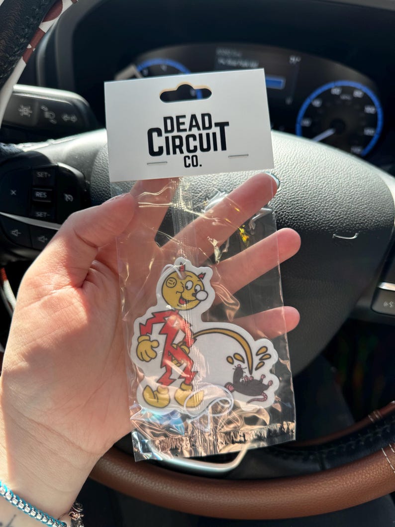 May include: A clear plastic package containing a sticker with a cartoon character in a red jumpsuit and a yellow face. The sticker also features a black rat and the text "DEAD CIRCUIT CO." on a white tag.