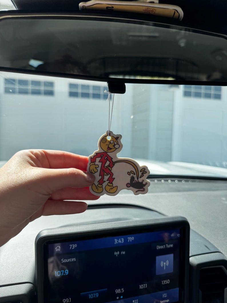 May include: A cartoon air freshener featuring a character in a red and yellow outfit, with a black mouse. The air freshener is hanging from a car's rearview mirror. The car's dashboard and radio display are visible in the foreground.