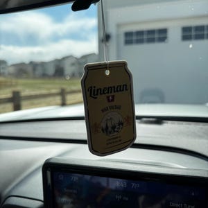 May include: A rectangular air freshener with the text "Lineman High Voltage" and a graphic of a power line tower and trees. The air freshener is hanging from a string in a car, with a view of a suburban neighborhood in the background.