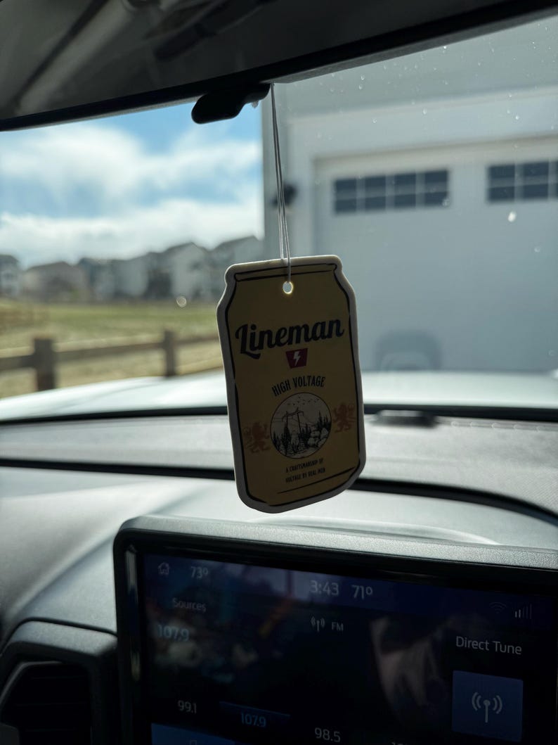 May include: A rectangular air freshener with the text "Lineman High Voltage" and a graphic of a power line tower and trees. The air freshener is hanging from a string in a car, with a view of a suburban neighborhood in the background.