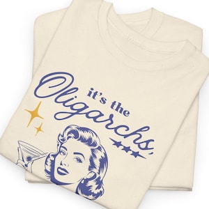 Puede incluir: Una camiseta beige con una ilustración azul y blanca de una mujer sosteniendo una copa de martini. El texto "it's the Oligarchs" está escrito en azul cursivo sobre la mujer.