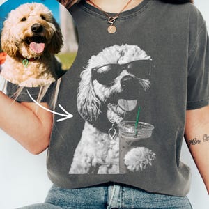 May include: A charcoal grey short-sleeve t-shirt featuring a black and white graphic of a curly-haired dog wearing sunglasses and holding a clear plastic cup with a green straw. A circular inset shows the original colour photograph of the dog.