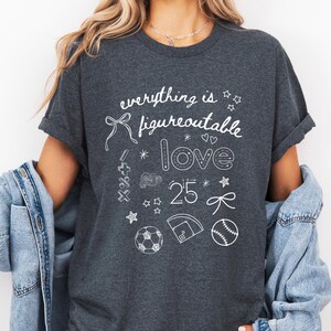 以下が含まれることがあります： 暗いグレーのTシャツに、白いグラフィックデザインが施されています。「everything is figureoutable love 25」という文字と、星、ハート、リボン、サッカーボール、野球ボール、野球場など、さまざまな落書きが描かれています。