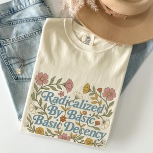 以下が含まれることがあります： クリーム色のTシャツ。青い文字で「Radicalized By Basic Basic Decency」と書かれ、ピンク、黄色、白の花のイラストで囲まれています。明るい色のジーンズ、ベージュの帽子、ネックレスと組み合わせています。