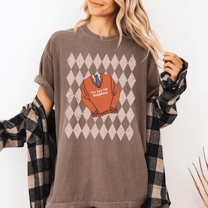 May include: Brown t-shirt with a graphic of an orange sweater and tie, set against a diamond pattern. The sweater graphic has the text "We Are All Neighbors". The shirt is a relaxed fit.