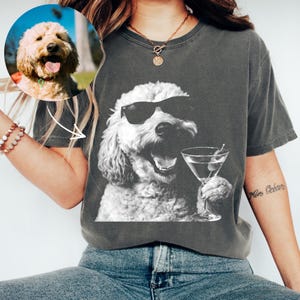 May include: A charcoal gray t-shirt featuring a black and white graphic print of a curly-haired dog wearing sunglasses and holding a martini glass. The dog has an open mouth and appears to be smiling. The shirt has a crew neckline and short sleeves.