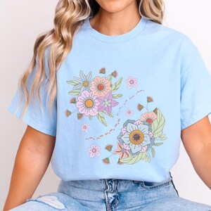 Könnte beinhalten: Ein hellblaues T-Shirt mit einem Blumendesign mit rosa, blauen, gelben und grünen Blumen. Die Blumen sind in einer Gruppe angeordnet, mit ein paar kleineren Blumen, die um sie herum verstreut sind.