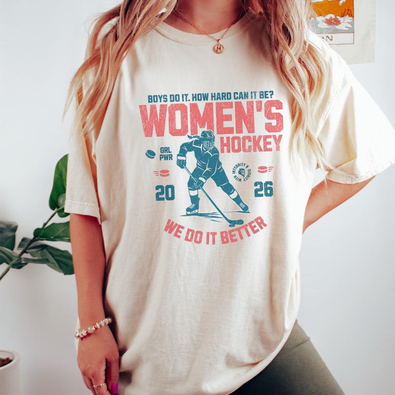 May include: Cream-colored t-shirt with a distressed graphic. The graphic reads "WOMEN'S HOCKEY" in red, with a teal hockey player illustration. Text includes "BOYS DO IT. HOW HARD CAN IT BE?" and "WE DO IT BETTER". Numbers "20" and "26" are also present.