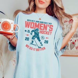 May include: Light blue t-shirt with the text "BOYS DO IT. HOW HARD CAN IT BE? WOMEN'S HOCKEY" in red. A teal hockey player graphic is centered below the text, with the words "WE DO IT BETTER" in red at the bottom.