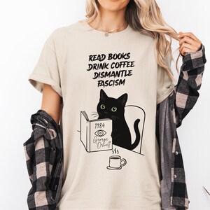 Könnte beinhalten: Beiges T-Shirt mit dem Text "READ BOOKS DRINK COFFEE DISMANTLE FASCISM". Eine schwarze Katze liest "1984" von George Orwell, darunter eine Kaffeetasse. Das T-Shirt hat einen Rundhalsausschnitt und kurze Ärmel.