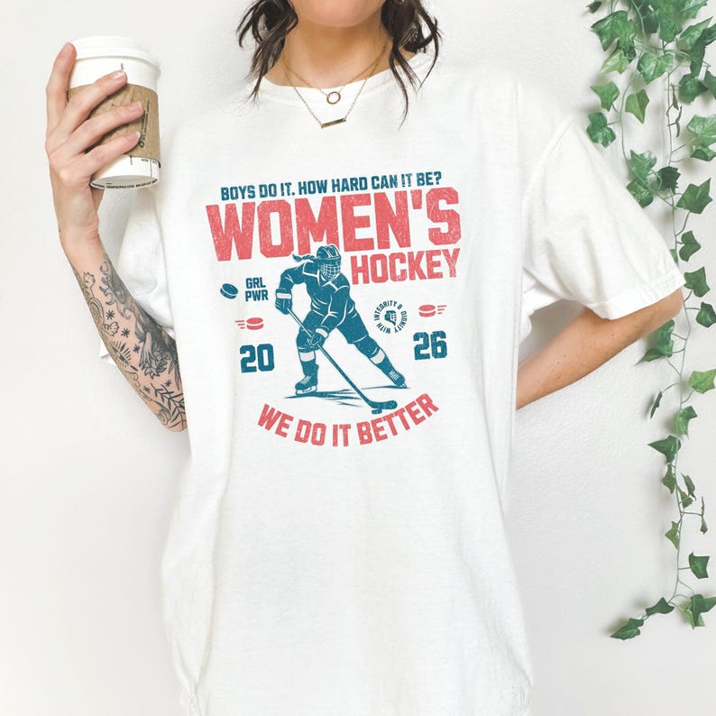 May include: White t-shirt with a hockey-themed graphic. The design features the text "WOMEN'S HOCKEY" in red, along with a hockey player illustration in blue. The shirt also includes the text "WE DO IT BETTER".