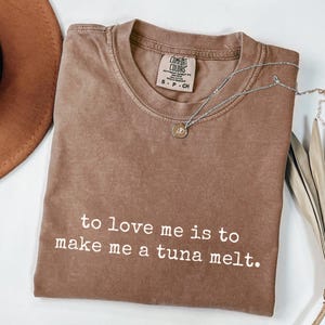 May include: A brown Comfort Colors t-shirt with the text "to love me is to make me a tuna melt" in white. A silver necklace with a small pendant is draped across the shirt. The shirt is folded on a white surface.
