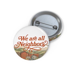 May include: A round pinback button featuring a landscape illustration of a small town with hills and the text We are all Neighbors in orange cursive script. The back of the button is silver metal with a horizontal pin mechanism attached.