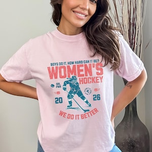 May include: A light pink t-shirt with the text "BOYS DO IT. HOW HARD CAN IT BE? WOMEN'S HOCKEY WE DO IT BETTER." A teal hockey player graphic is in the center. The shirt also has the numbers "20" and "26".