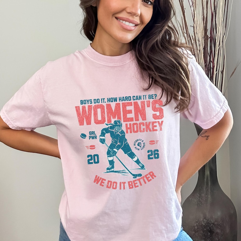 May include: A light pink t-shirt with the text "BOYS DO IT. HOW HARD CAN IT BE? WOMEN'S HOCKEY WE DO IT BETTER." A teal hockey player graphic is in the center. The shirt also has the numbers "20" and "26".