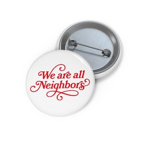 Op de afbeelding: Een ronde witte button met de rode tekst We are all Neighbors in een cursief lettertype. De achterkant van de button is van zilverkleurig metaal met een horizontale speldbevestiging om aan kleding of stoffen voorwerpen te bevestigen.