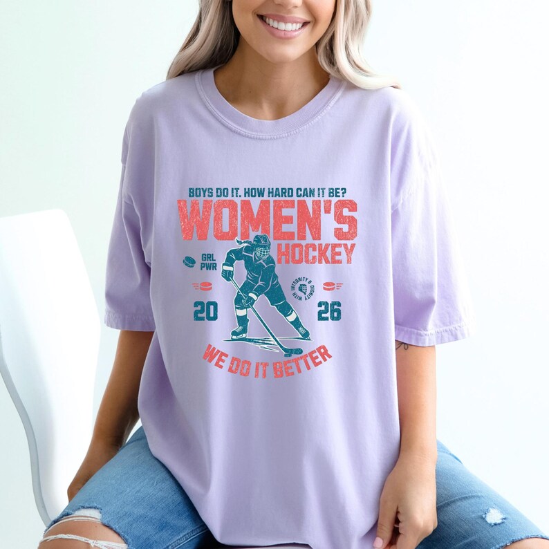 May include: A light purple t-shirt with the text "WOMEN'S HOCKEY" in red and teal. The shirt features a hockey player graphic and the words "WE DO IT BETTER". The numbers 20 and 26 are also displayed.