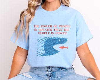 May include: Light blue t-shirt with the text "THE POWER OF PEOPLE IS GREATER THAN THE PEOPLE IN POWER" in red. The graphic shows a large blue fish eating a small red fish, with a school of blue fish.