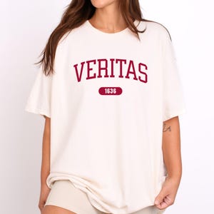 以下が含まれることがあります： 赤い「VERITAS」のグラフィックと下に印刷された「1636」の年が書かれた白いTシャツです。