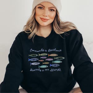 May include: Black sweatshirt with the text "Community is Resistance" and "Diversity is our Strength." The sweatshirt features a colorful fish design. The model is wearing a beige beanie.