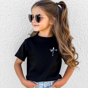 K&ouml;nnte beinhalten: Ein junges M&auml;dchen tr&auml;gt ein schwarzes T-Shirt mit einem wei&szlig;en Kreuz und dem Text "JOHN 3:16" darauf. Sie tr&auml;gt herzf&ouml;rmige Sonnenbrillen und hat ihre Haare zu einem Pferdeschwanz gebunden.